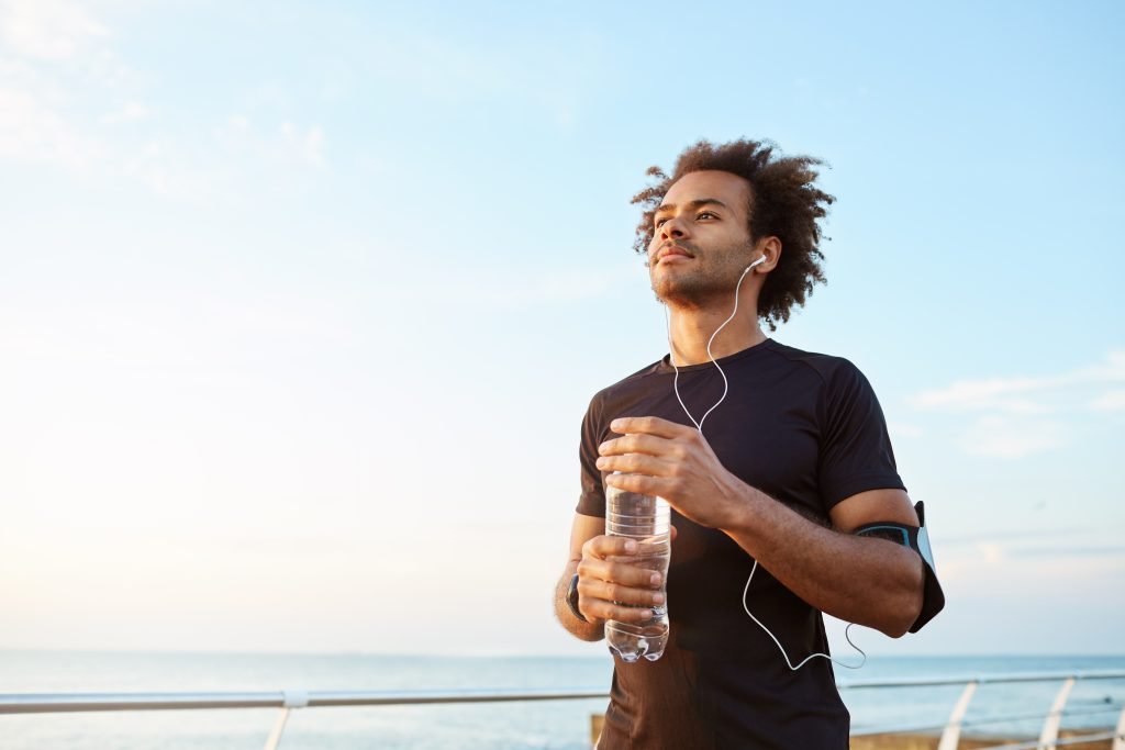 Homem vestindo roupa esportiva preta e fones de ouvido, segurando uma garrafa de água e olhando para o horizonte com o mar ao fundo.