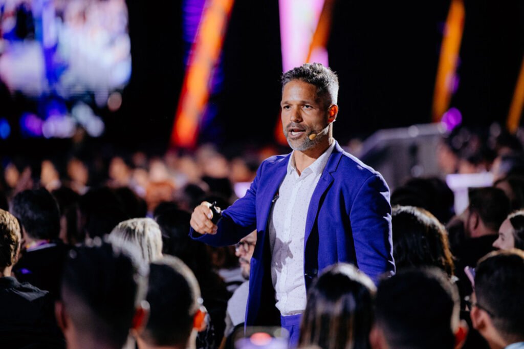 Fotografia em plano médio de um palestrante (Diego Ribas) visto de perfil, vestindo paletó azul e camisa branca. Ele segura um passador de slides e fala ao microfone headset para uma plateia lotada e desfocada em um ambiente de evento. A iluminação é cênica, com pontos de luz coloridos ao fundo.