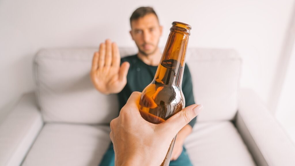 Fotografia em primeira pessoa mostrando uma mão oferecendo uma garrafa de cerveja de vidro castanho. Ao fundo, um homem sentado em um sofá branco faz um gesto de "pare" com a mão aberta, recusando a bebida. O homem está levemente fora de foco, dando ênfase ao gesto de negação.