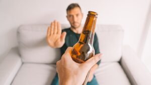 Fotografia em primeira pessoa mostrando uma mão oferecendo uma garrafa de cerveja de vidro. Ao fundo, um homem sentado em um sofá branco faz um gesto de "pare" com a mão aberta, recusando a bebida. O homem está levemente fora de foco, dando ênfase ao gesto de negação.
