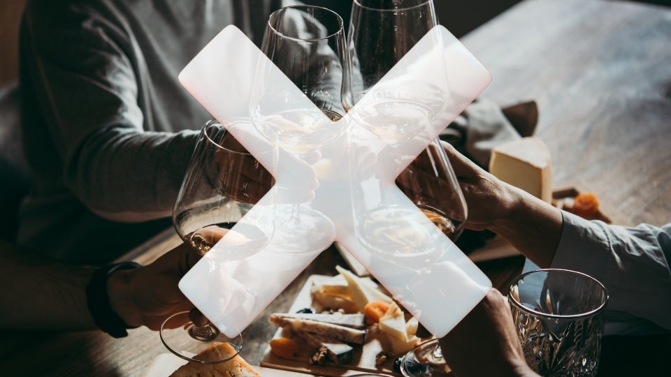 Quatro pessoas brindam com taças de vinho e copos de cristal sobre uma mesa com tábuas de queijos e frutas secas. Sobreposta à imagem, há uma grande marca de "X" branca e translúcida, simbolizando a proibição ou a recusa ao consumo de bebidas alcoólicas naquele contexto.