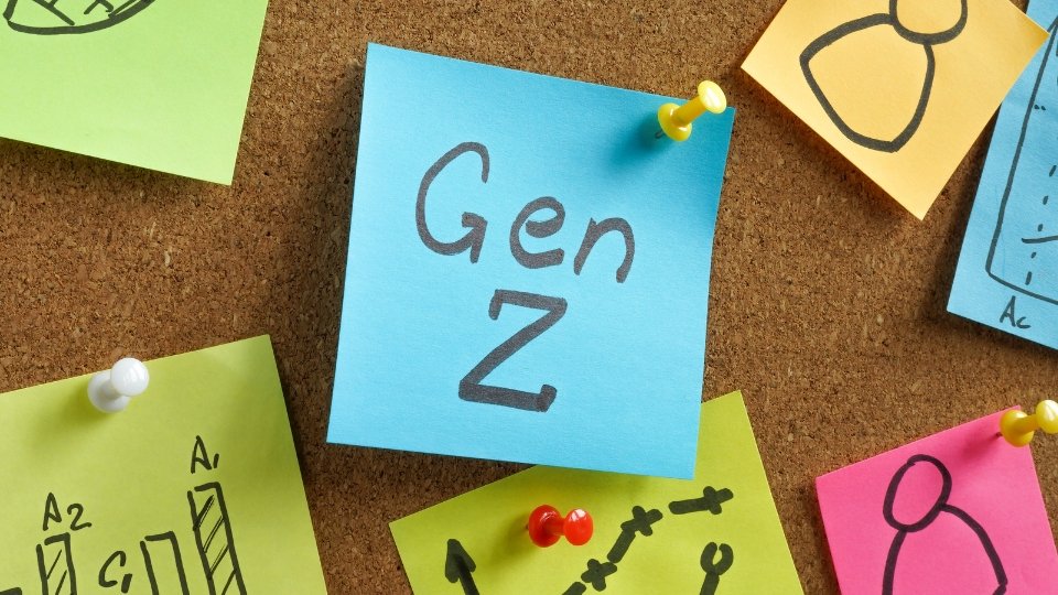Close-up de um mural de cortiça com vários papéis adesivos coloridos. No centro, um post-it azul claro tem a escrita a palavra "Gen Z" 