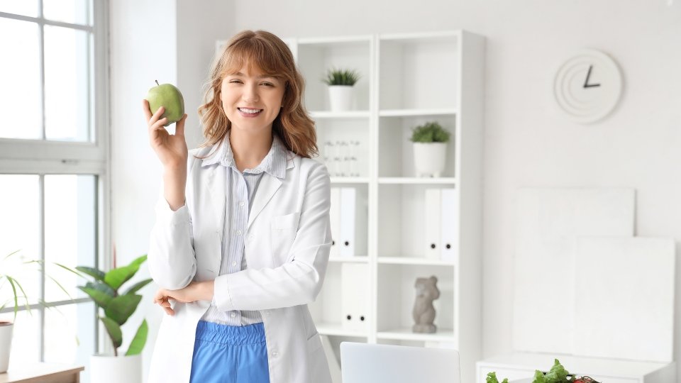 Nutricionista sorridente vestindo jaleco branco e segurando uma maçã verde em um consultório claro e moderno.