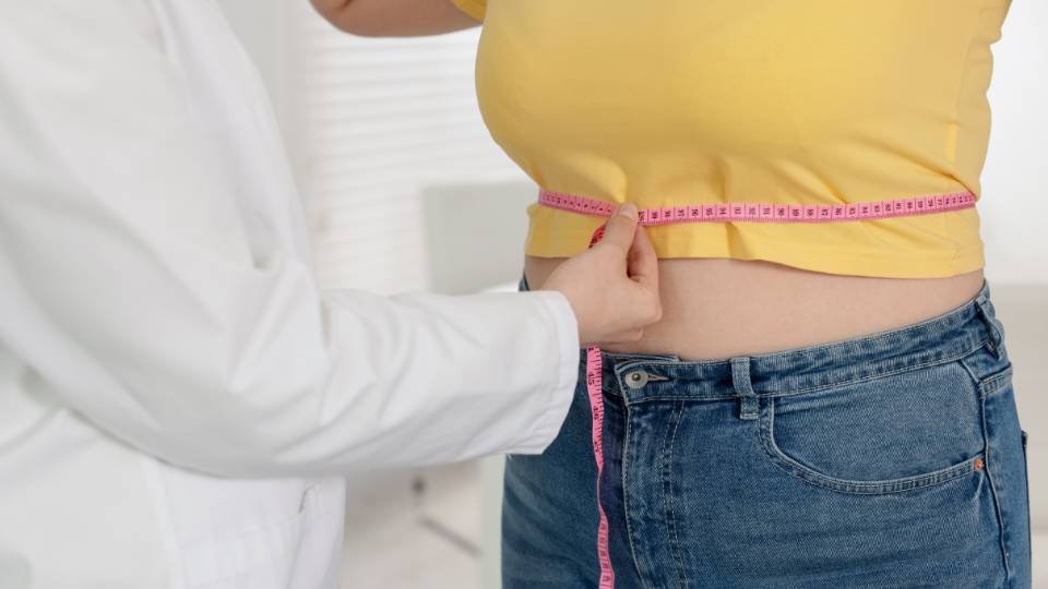 Close-up lateral de um profissional de saúde usando uma fita métrica rosa para medir a cintura de uma pessoa que veste uma camiseta amarela e calça jeans. A imagem foca no monitoramento de medidas corporais em um ambiente clínico.