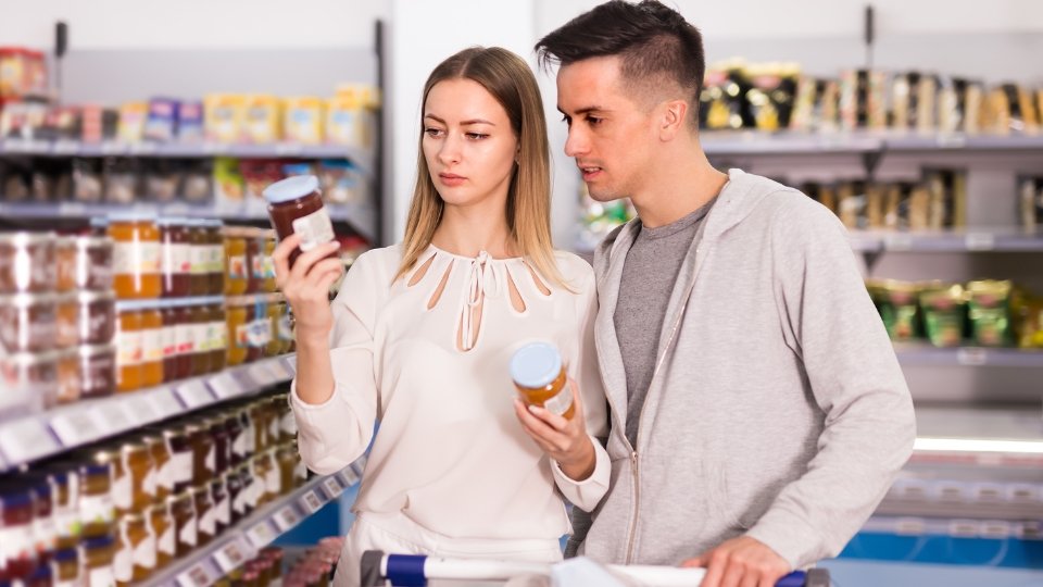 Casal no corredor de um supermercado lendo atentamente as informações nutricionais do rótulo de um produto em embalagem de vidro.