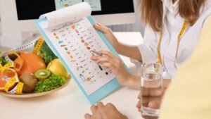 ista superior de uma consulta nutricional. Uma profissional de jaleco aponta com uma caneta para uma tabela de alimentos ilustrada em uma prancheta. Sobre a mesa, há uma cesta com frutas frescas (kiwi, maçã e laranja), uma fita métrica e um copo d'água.