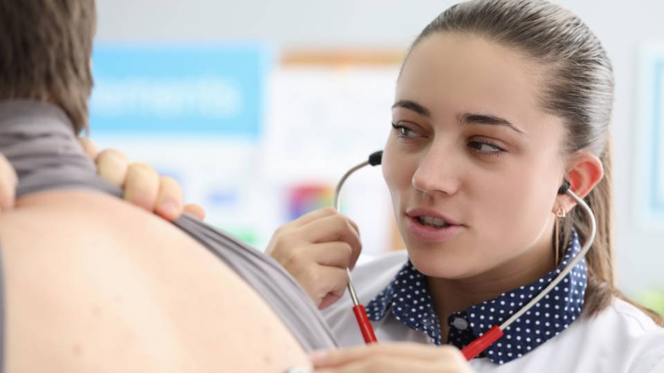 Médica de jaleco branco e camisa de poá utilizando um estetoscópio vermelho para realizar um exame clínico nas costas de um paciente.