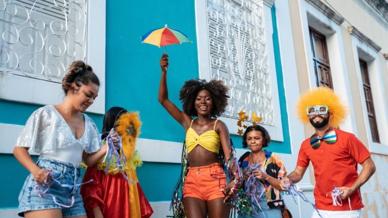 Um grupo alegre de foliões celebra o Carnaval em frente a uma fachada azul vibrante com janelas brancas de estilo colonial. No centro, uma mulher negra sorridente segura uma pequena sombrinha de frevo colorida, vestindo um top amarelo e shorts laranja com adereços brilhantes. À direita, um homem usa uma peruca afro amarela, óculos azuis gigantes e gravata borboleta colorida. O grupo se diverte com serpentinas e fantasias criativas, capturando a energia vibrante de um bloco de rua.