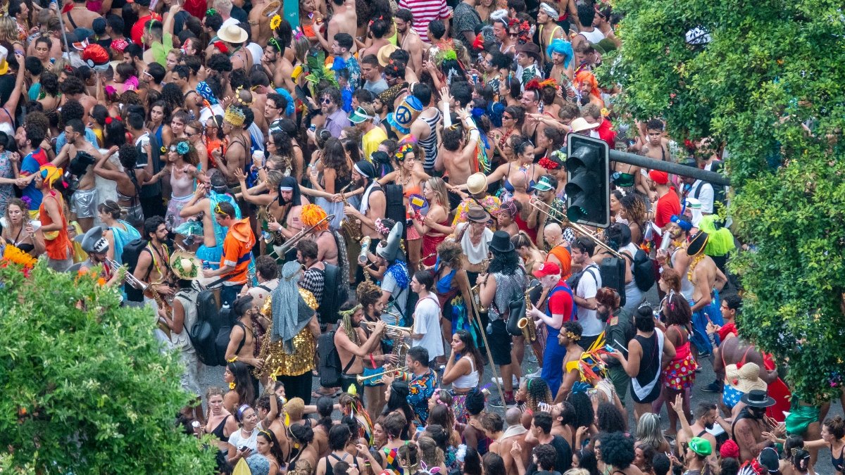 Vista aérea de um bloco de Carnaval de rua densamente povoado. A multidão, vestindo fantasias coloridas e brilhantes, preenche toda a extensão da rua entre árvores verdes. É possível identificar diversos músicos com instrumentos de sopro, como trompetes e saxofones, em meio aos foliões, capturando a energia e a escala monumental da festa popular.