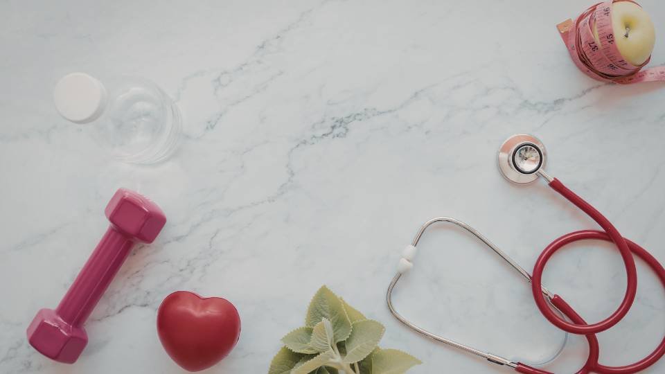 Visão superior sobre fundo de mármore com itens de saúde: um estetoscópio vermelho, um haltere rosa, um coração decorativo e uma fita métrica envolvendo uma maçã.