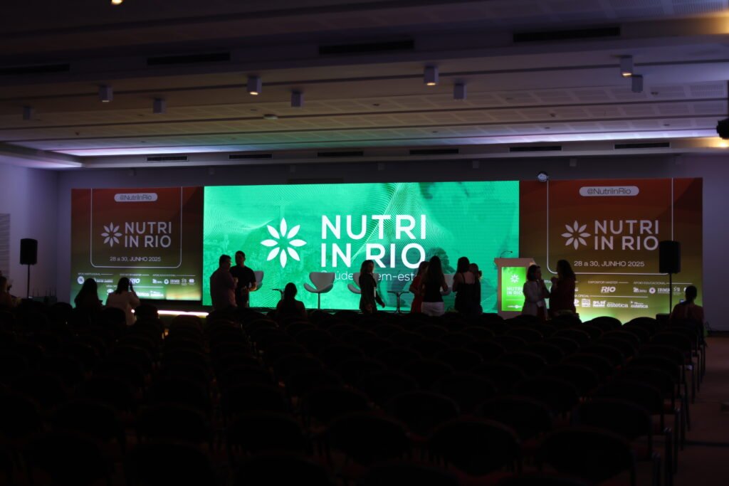 Auditório do evento Nutri In Rio com participantes interagindo em frente ao palco principal, que exibe um grande painel de LED verde com a logo do congresso.