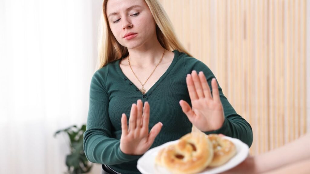 Mulher fazendo gesto de recusa com as mãos para um prato com pães oferecido a ela, ilustrando restrição alimentar.
