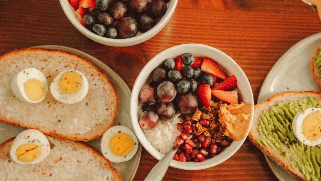 Mesa de café da manhã saudável vista de cima com bowls de iogurte, frutas, granola e torradas com abacate e ovo cozido.