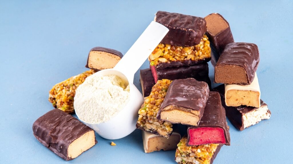 Várias barras de proteína cortadas ao meio, revelando diferentes recheios como caramelo, amendoim e uma opção com interior rosa, estão empilhadas sobre um fundo azul claro. À esquerda da pilha, um medidor plástico branco (scoop) aparece cheio de suplemento em pó.