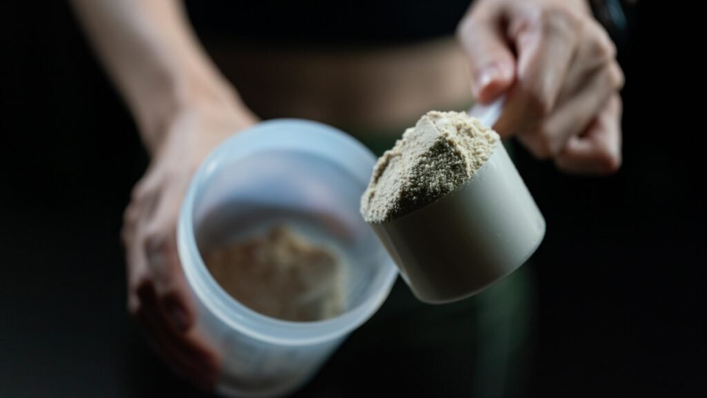 Imagem em plano detalhe e contra a luz mostrando as mãos de uma pessoa preparando um suplemento, um whey protein. Uma das mãos segura uma coqueteleira plástica aberta, enquanto a outra segura um medidor cheio de suplemento em pó, prestes a despejá-lo no recipiente. O fundo é escuro.