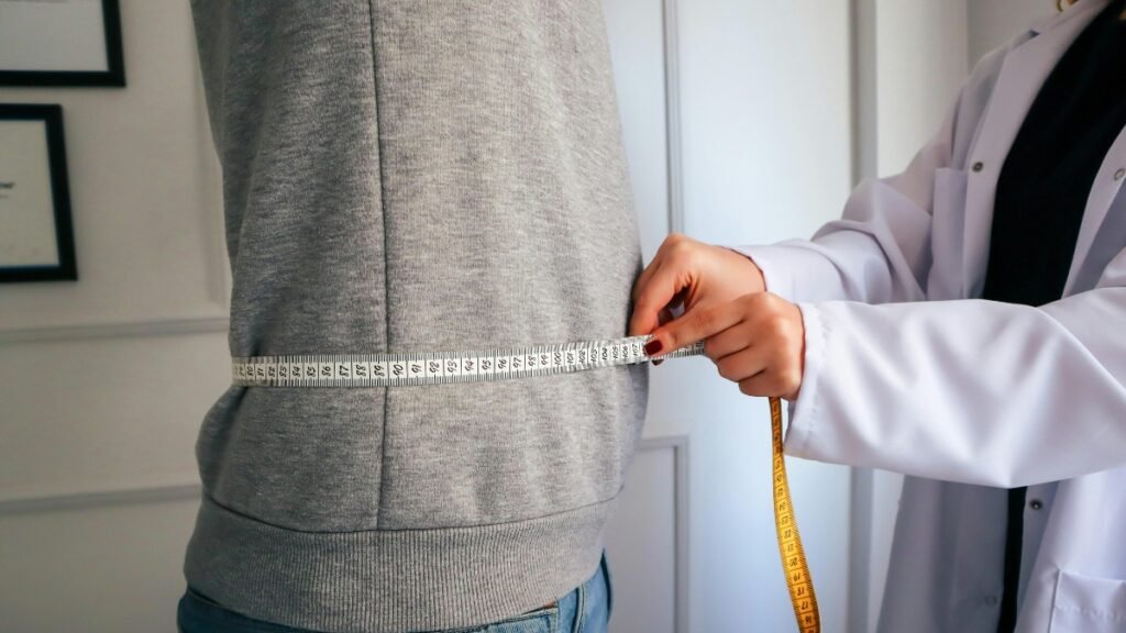 Close-up lateral de um profissional de saúde vestindo jaleco branco enquanto usa uma fita métrica branca e amarela para medir a circunferência abdominal de um paciente. O paciente está de perfil, vestindo um moletom cinza e calça jeans. Ao fundo, a parede branca exibe quadros com moldura preta.