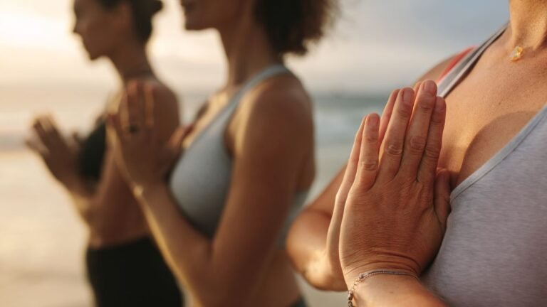 Close-up lateral de pessoas alinhadas praticando meditação ou yoga na praia durante o amanhecer ou pôr do sol. Em destaque e em primeiro plano, uma mulher aparece com as mãos unidas em posição de prece na altura do peito. As outras pessoas ao fundo seguem a mesma postura, aparecendo progressivamente desfocadas.