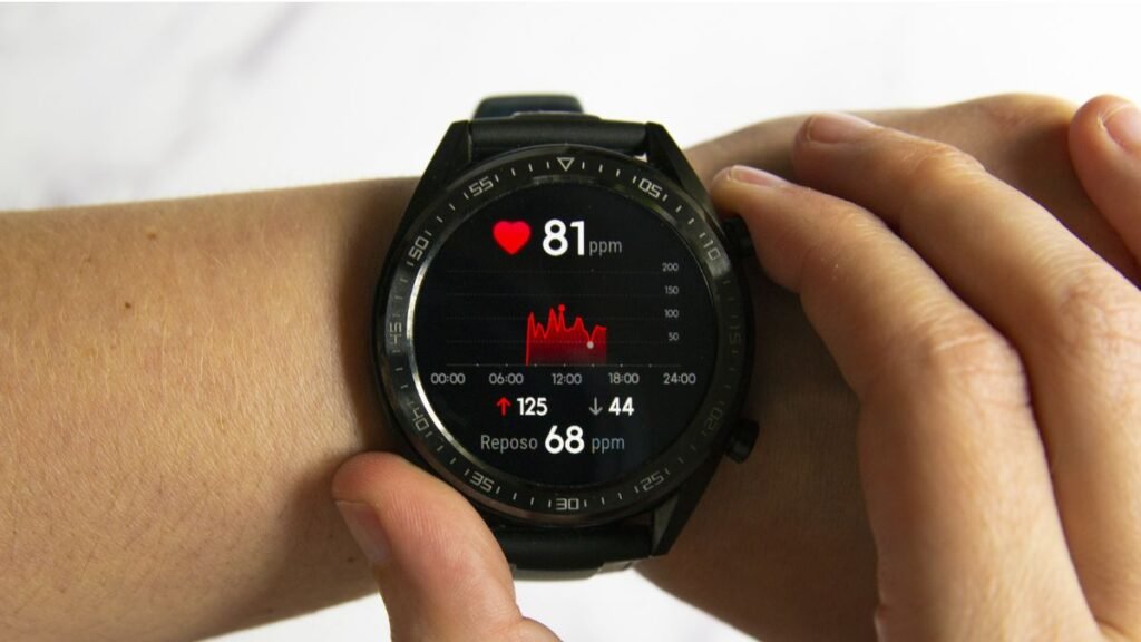 Close-up de um smartwatch preto colocado no pulso de uma pessoa, cujo mostrador exibe dados de monitoramento cardíaco. Na tela, lê-se "81 ppm" ao lado de um coração vermelho, acompanhado por um gráfico de linha e os indicadores "125", "44" e "Reposo 68 ppm". Os dedos da outra mão tocam a borda do relógio.