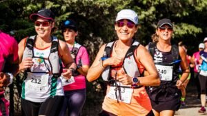 Grupo de mulheres sorridentes participa de uma corrida de trilha durante o dia. Em primeiro plano, uma corredora ao centro veste camiseta laranja, boné e óculos escuros, exibindo o número de peito "443" e uma mochila de hidratação. Ao seu lado esquerdo, outra corredora usa blusa branca com detalhes verdes e pretos e o número "232". Outras atletas com roupas esportivas, números de inscrição e óculos escuros correm logo atrás, cercadas por árvores e vegetação nativa ao fundo.