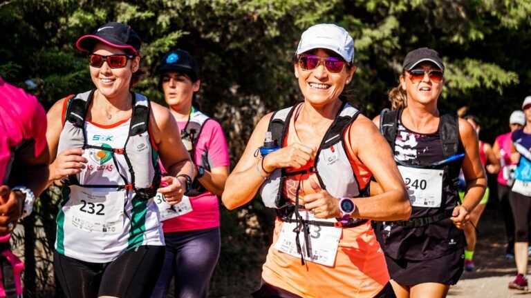Grupo de mulheres sorridentes participa de uma corrida de trilha durante o dia. Em primeiro plano, uma corredora ao centro veste camiseta laranja, boné e óculos escuros, exibindo o número de peito "443" e uma mochila de hidratação. Ao seu lado esquerdo, outra corredora usa blusa branca com detalhes verdes e pretos e o número "232". Outras atletas com roupas esportivas, números de inscrição e óculos escuros correm logo atrás, cercadas por árvores e vegetação nativa ao fundo.