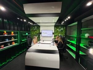 Aqui está o alt text para esta imagem: Alt Text: Vista ampla de um estúdio moderno de gravação de podcast e gravação de vídeo, iluminado com luzes de LED verdes. Três mulheres estão sentadas ao redor de uma grande mesa branca e curvada com microfones profissionais. Ao fundo, uma parece decorada com plantas artificiais abriga uma televisão com o logotipo "Life Global". Nas laterais, há estantes pretas com detalhes iluminados, exibindo colecionáveis de personagens da cultura pop e equipamentos.