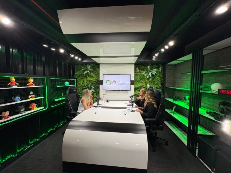 Aqui está o alt text para esta imagem: Alt Text: Vista ampla de um estúdio moderno de gravação de podcast e gravação de vídeo, iluminado com luzes de LED verdes. Três mulheres estão sentadas ao redor de uma grande mesa branca e curvada com microfones profissionais. Ao fundo, uma parece decorada com plantas artificiais abriga uma televisão com o logotipo "Life Global". Nas laterais, há estantes pretas com detalhes iluminados, exibindo colecionáveis de personagens da cultura pop e equipamentos.
