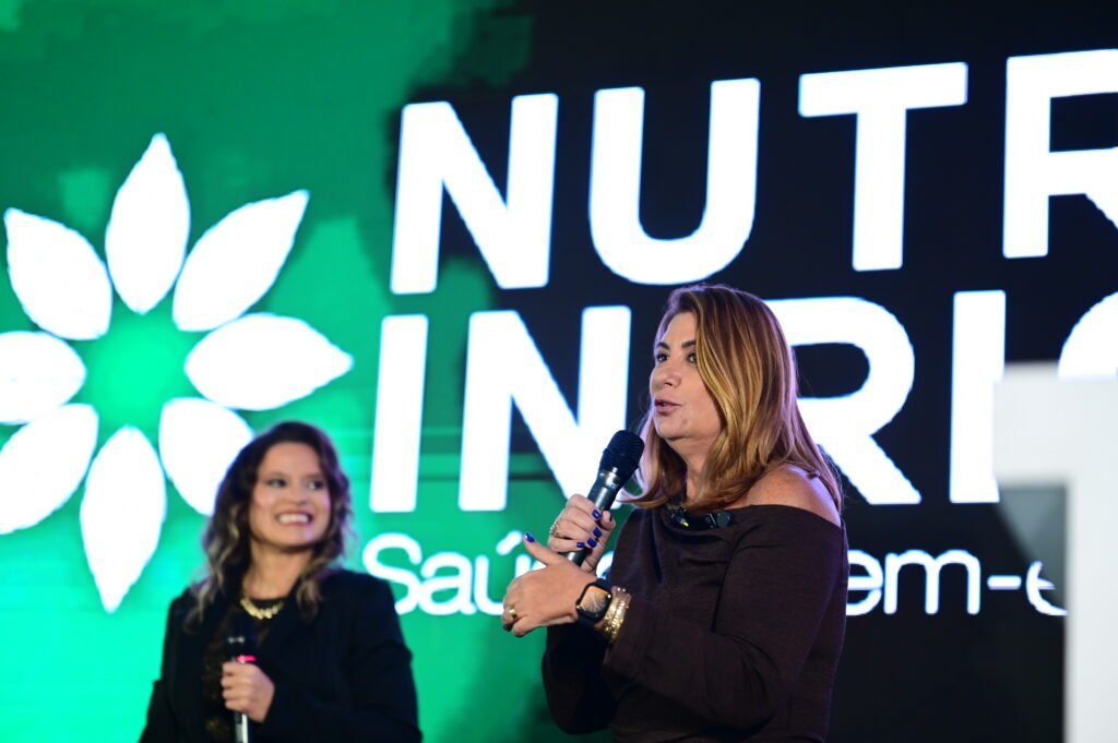 Fotografia em plano médio capturada durante um evento no palco. Em primeiro plano, à direita, uma mulher loira de ombros de fora fala ao microfone. Ela gesticula com a mão esquerda e usa pulseiras e um relógio. Ao fundo, à esquerda, outra mulher sorri enquanto segura seu próprio microfone. Atrás delas, um grande painel verde e preto exibe o logotipo "NUTRI RIO" em letras brancas grandes, com o subtítulo "Saúde e bem-estar".
