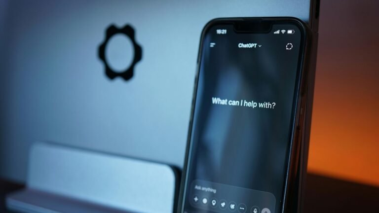 Close-up de um smartphone em pé exibindo a aplicativo do ChatGPT em modo escuro na tela, com a pergunta "What can I help with?" no centro. Ao fundo, levemente desfocado, há um computador prateado e uma iluminação com tom alaranjado à direita.