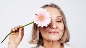 Retrato em close-up de uma mulher madura de cabelos grisalhos curtos e sorriso suave. Ela segura o caule de uma gérbera rosa claro, cobrindo delicadamente um dos olhos com a flor. O fundo é neutro e bem iluminado.