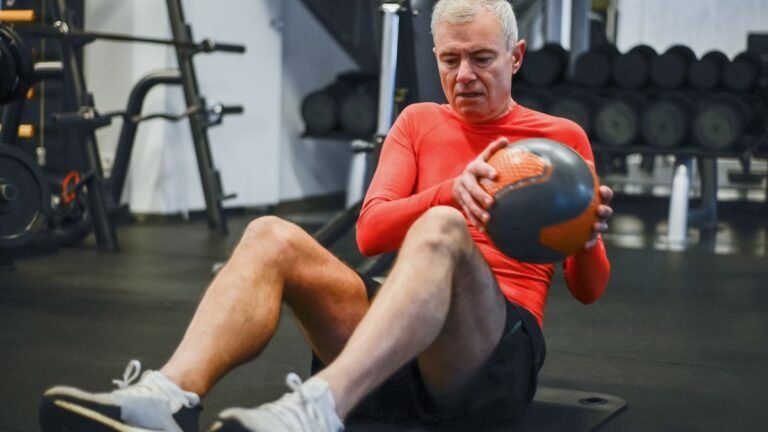 Homem de meia-idade com cabelos grisalhos e camisa esportiva vermelha de mangas compridas realiza abdominal com rotação (Russian twist) em uma academia. Ele está sentado sobre um colchonete no chão, com os joelhos dobrados, segurando uma medicina ball cinza e laranja com as duas mãos perto do tronco. Ao fundo, veem-se aparelhos de musculação e halteres.