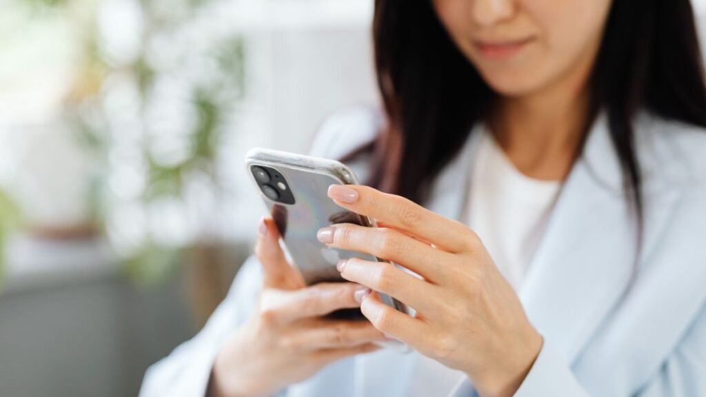 Plano detalhe focado nas mãos de uma mulher com unhas pintadas de bege, segurando e digitando em um smartphone cinza com capinha transparente. Ela veste blazer azul-claro sobre camiseta branca, com o rosto parcialmente visível e levemente desfocado em segundo plano.
