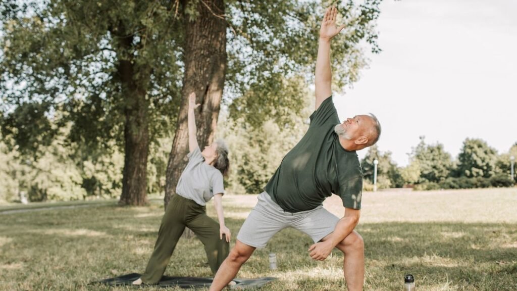 Casal idoso pratica yoga em um gramado ensolarado ao ar livre, cercado por árvores. Em primeiro plano, o homem de barba grisalha, camiseta verde e shorts cinza realiza a postura do ângulo lateral estendido, apoiando um antebraço na coxa dobrada e elevando o outro braço em direção ao céu. Ao fundo, uma mulher realiza a mesma postura sobre um tapete de yoga.