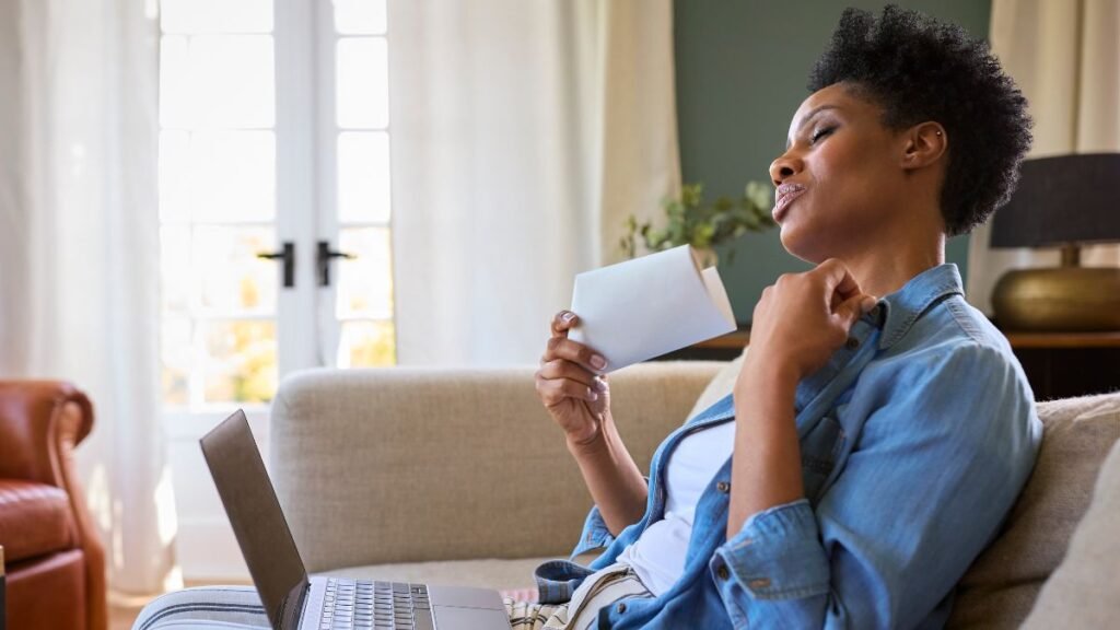 Mulher de cabelo curto vestindo camisa jeans e blusa branca, sentada em um sofá claro com um laptop aberto ao lado. Ela se abana com um bloco de papéis dobrado e puxa a gola da camisa, expressando desconforto com o calor ou calor das ondas da menopausa.