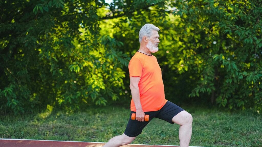 Homem idoso de cabelos e barba brancos realiza o exercício de afundo (lunge) de perfil em uma pista ao ar livre. Ele veste camiseta laranja vibrante e shorts preto, segurando um pequeno halter laranja em uma das mãos estendidas ao lado do corpo. O fundo conta com folhagens e árvores bem iluminadas pela luz natural.