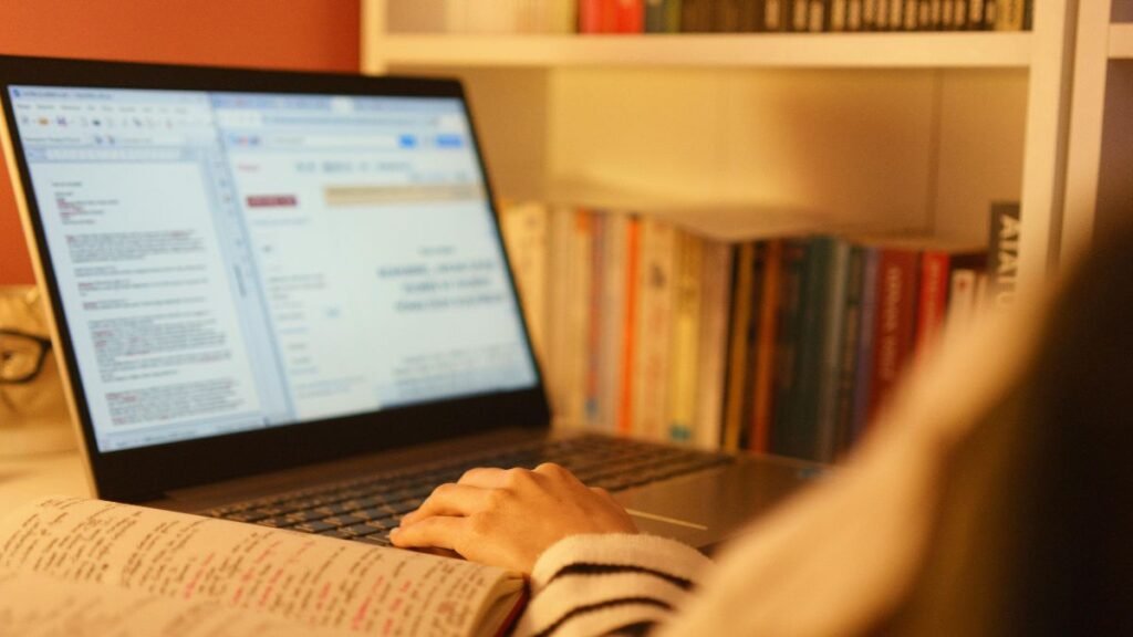 Visão por cima do ombro de uma pessoa digitando no touchpad de um laptop aberto em uma escrivaninha iluminada por luz quente. Na tela, vê-se um documento de texto e uma página web abertos. Em primeiro plano, há um caderno com anotações e, ao fundo, uma estante com livros coloridos.